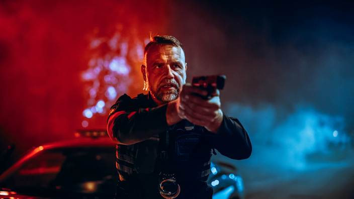 Police officer in tactical gear points a gun, with police car lights flashing. The image is associated with content about wrongful police shootings during raids.